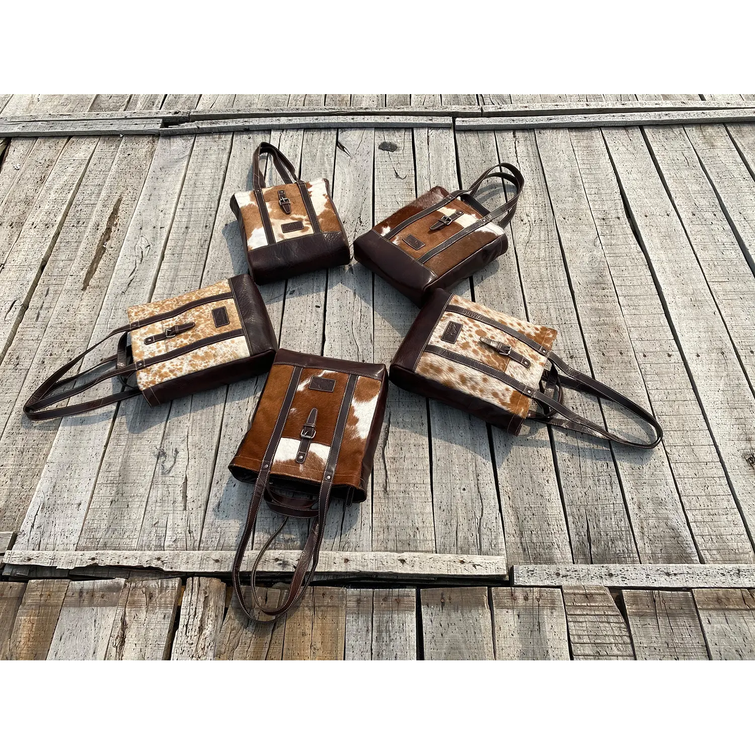 Fünf Shopper-HALLODRI-Taschen mit Kuhfellmuster und braunen Griffen sind in Form eines kreisförmigen Sterns auf einer rustikalen Holzoberfläche angeordnet und verfügen jeweils über einzigartige Flicken für eine symmetrische Präsentation.