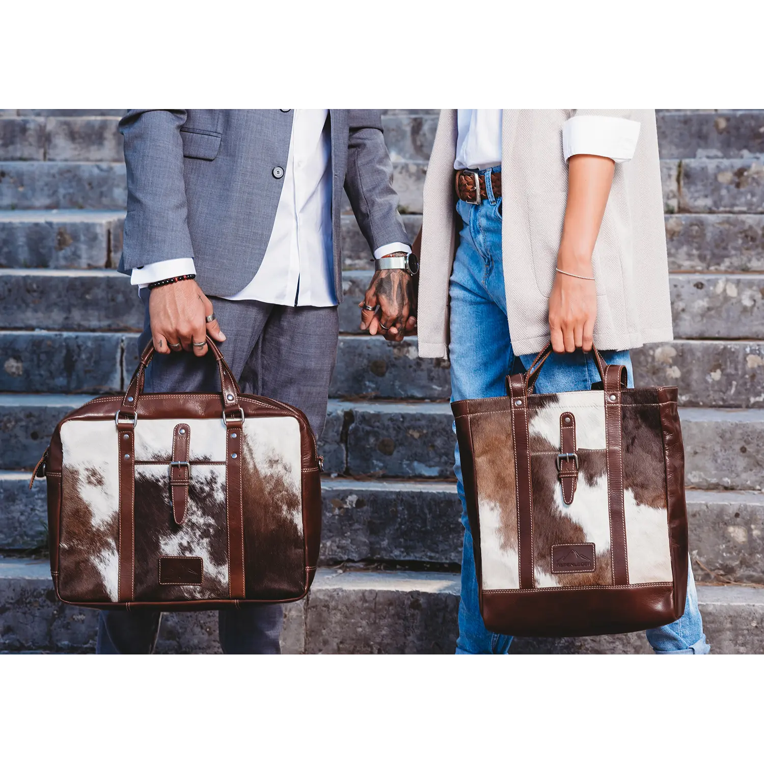 Zwei Personen halten sich auf einer Steintreppe an den Händen und tragen jeweils einen stylischen Alpenleder Shopper HALLODRI aus einzigartigem braun-weißem Leder. Die linke Person trägt einen grauen Anzug, die rechte einen hellen Mantel und Jeans.