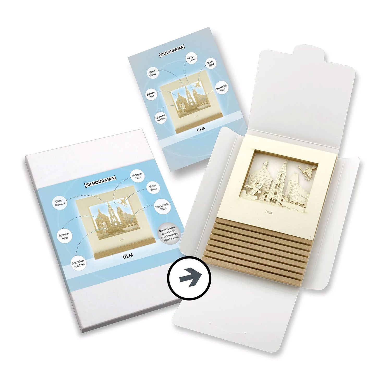 Eine geöffnete weiße Ulm-SILHOURAMA Silhoubox L enthält eine 3D-Stadtansicht mit übereinanderliegenden Papierausschnitten eines Münsters mit dem Titel „Ulm“ sowie ein Montageheft. Die mit blauen Akzenten und kreisförmigen Illustrationen verzierte Verpackung macht sie zum idealen Geschenk für Ulm-Fans.