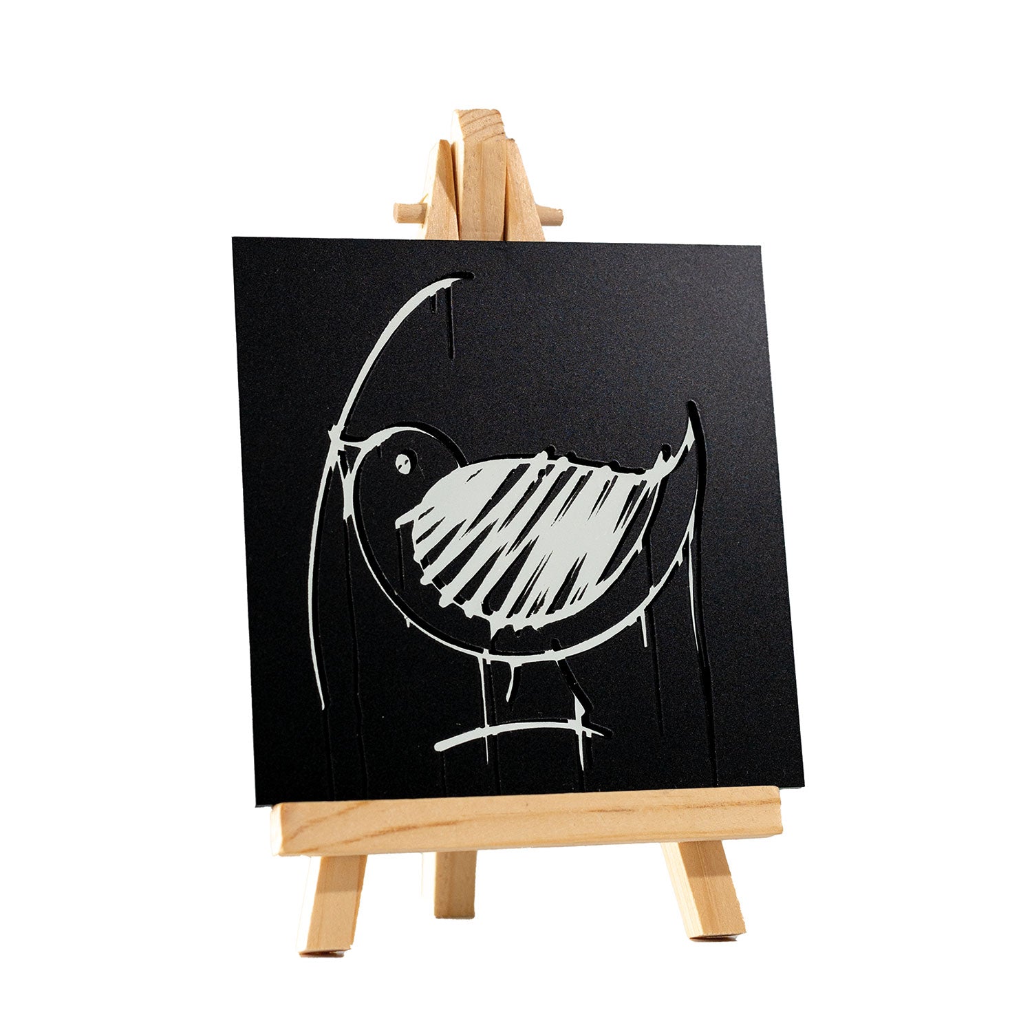 Ein kleiner hölzerner Ulmer Spatz Designspiegel Staffelei zeigt eine schwarze quadratische Leinwand mit einer minimalistischen, weißen, kreideartigen Vogelskizze mit kräftigen Linien und kreisförmigen Formen vor einem schlichten Hintergrund.