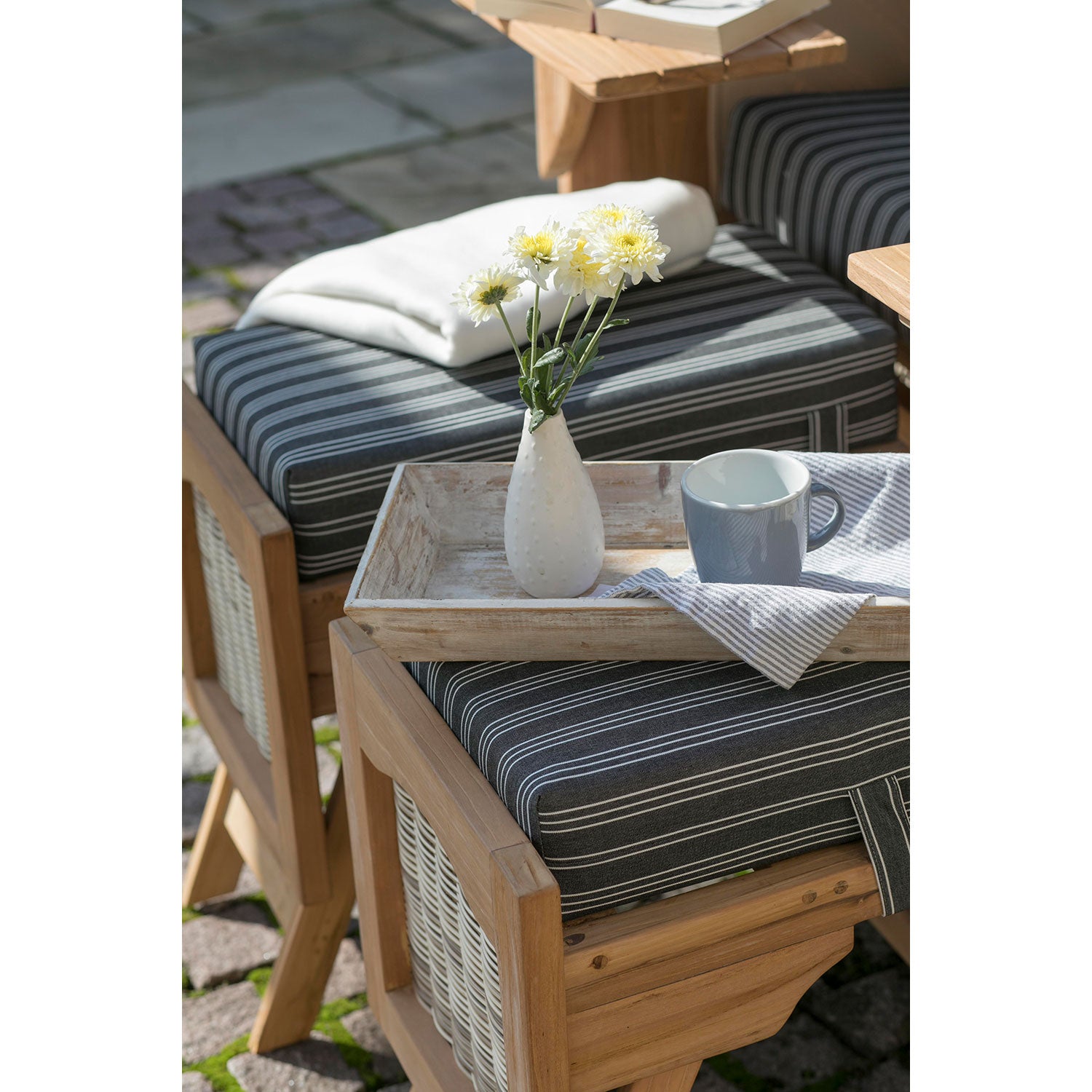 Der Strandkorb DÜNE, grau mit Nadelstreifen, besteht aus einem Tablett mit einer weißen Vase mit gelben und weißen Blumen, einer blauen Tasse und einer gestreiften Serviette. Eine gefaltete weiße Decke unterstreicht die Gemütlichkeit dieses eleganten Ambientes.