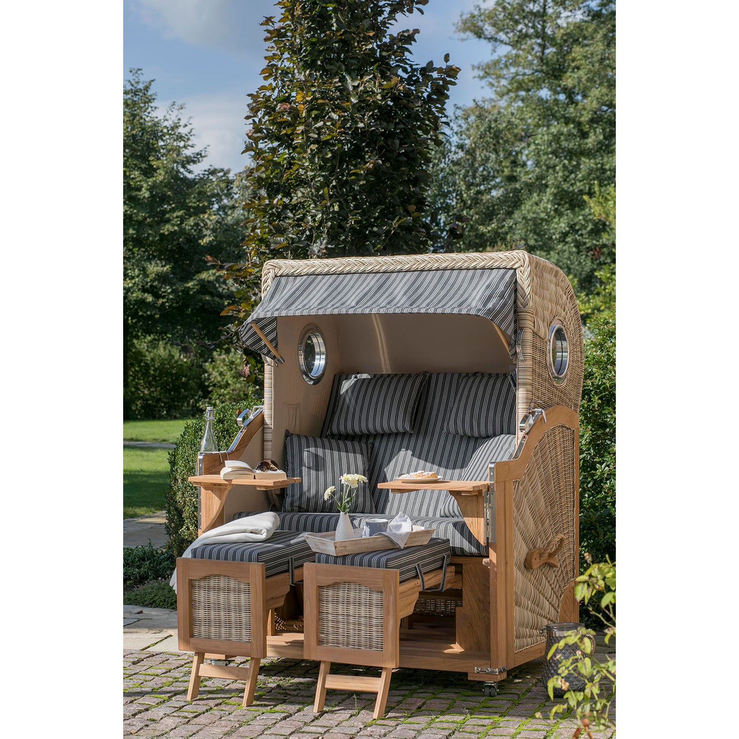 Der Strandkorb DÜNE, grau mit Nadelstreifen, steht auf einer Steinterrasse im Garten. Er verfügt über verstellbare Fußstützen, integrierte Getränkeablagen und eine charmante Pflanzendekoration. Bäume und Grünflächen bilden eine ruhige Kulisse.