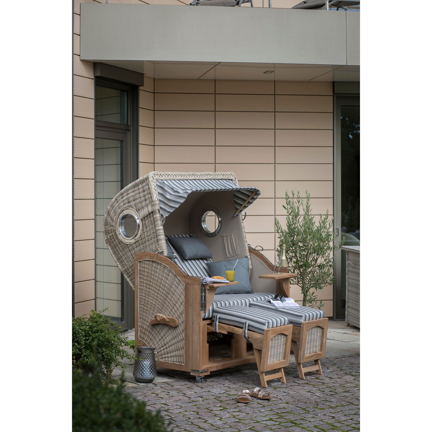 Ein Strandkorb DÜNE, grau gestreift, mit Fußstützen und einem gewölbten Baldachin mit runden Fenstern steht auf der Terrasse eines modernen Gebäudes. Gefertigt aus PE-Geflecht, wird er von einem Olivenbaum und einem kleinen Tisch mit Tellern und einer Tasse begleitet und schafft so eine ruhige Atmosphäre.