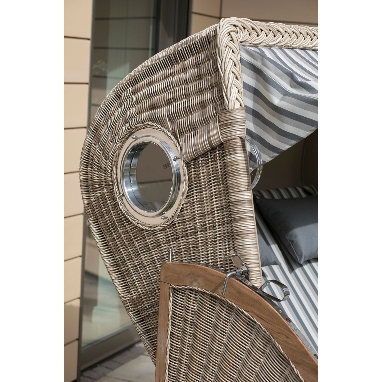 Nahaufnahme des Strandkorbs DÜNE, eines grau gestreiften Strandkorbs aus Teakholz mit gestreiften Kissen und einem runden Fenster mit poliertem Metallrahmen in der Korbseitenwand. Im Hintergrund harmoniert eine beige geflieste Gebäudefassade mit dem eleganten Design.