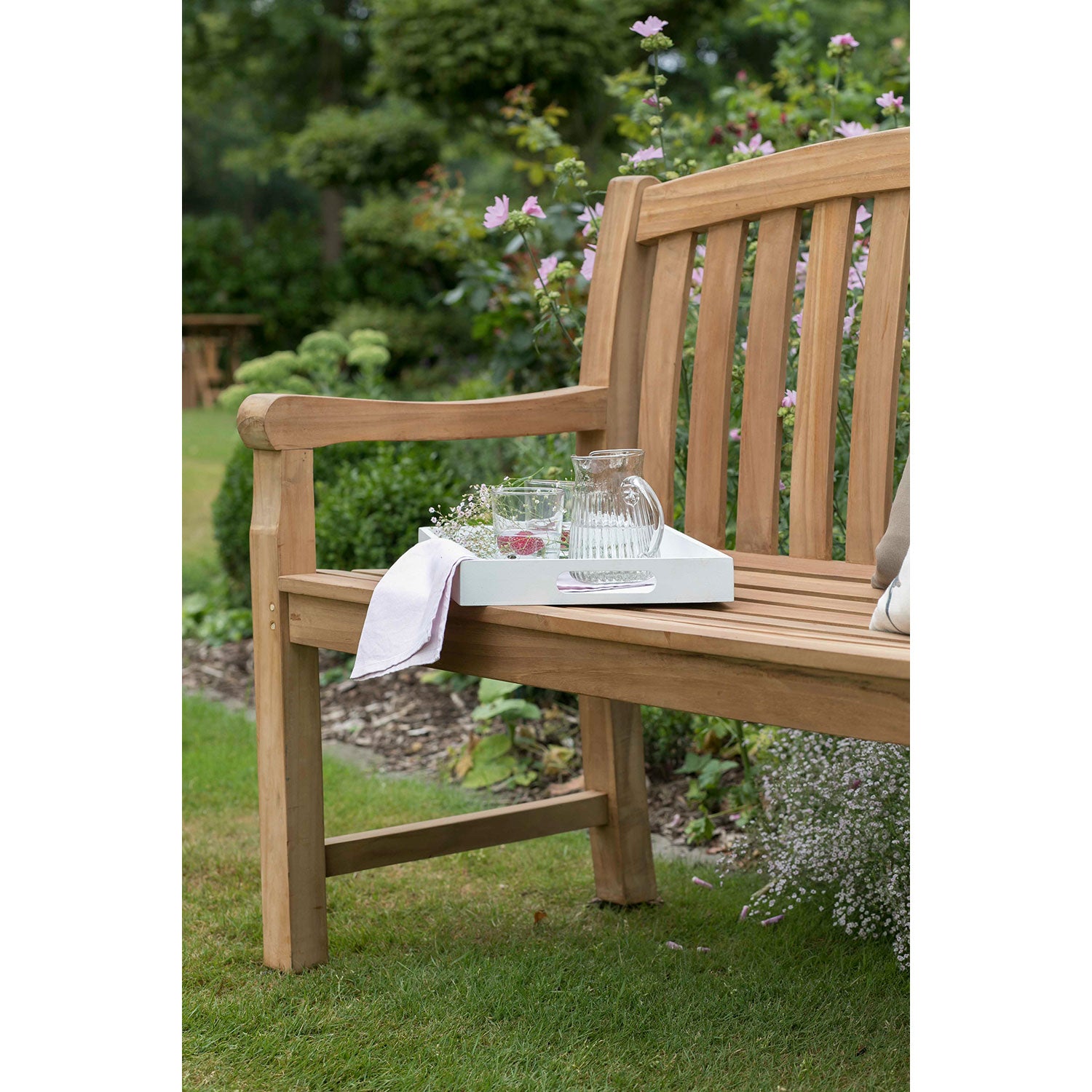 Die Gartenbank aus Holz 2-Sitzer wird mit einem weißen Tablett, einem Glaskrug, zwei Gläsern und einer Serviette zum stilvollen Highlight für die Outdoor-Gestaltung zwischen grünem Rasen, Blumen und Sträuchern.