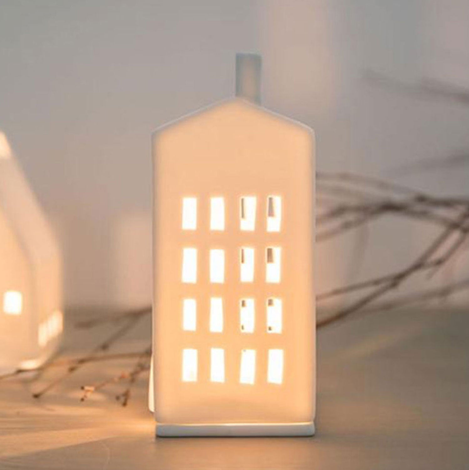 Das Lichthaus „Mittel, Kleine Fenster“ ist eine handgefertigte Porzellanlampe in Hausform mit mehreren kleinen Fenstern und einem Kamin. Sie strahlt warmes Licht aus und schafft eine gemütliche Atmosphäre, perfekt für die Weihnachtsdekoration mit verschwommenen Zweigen im Hintergrund.