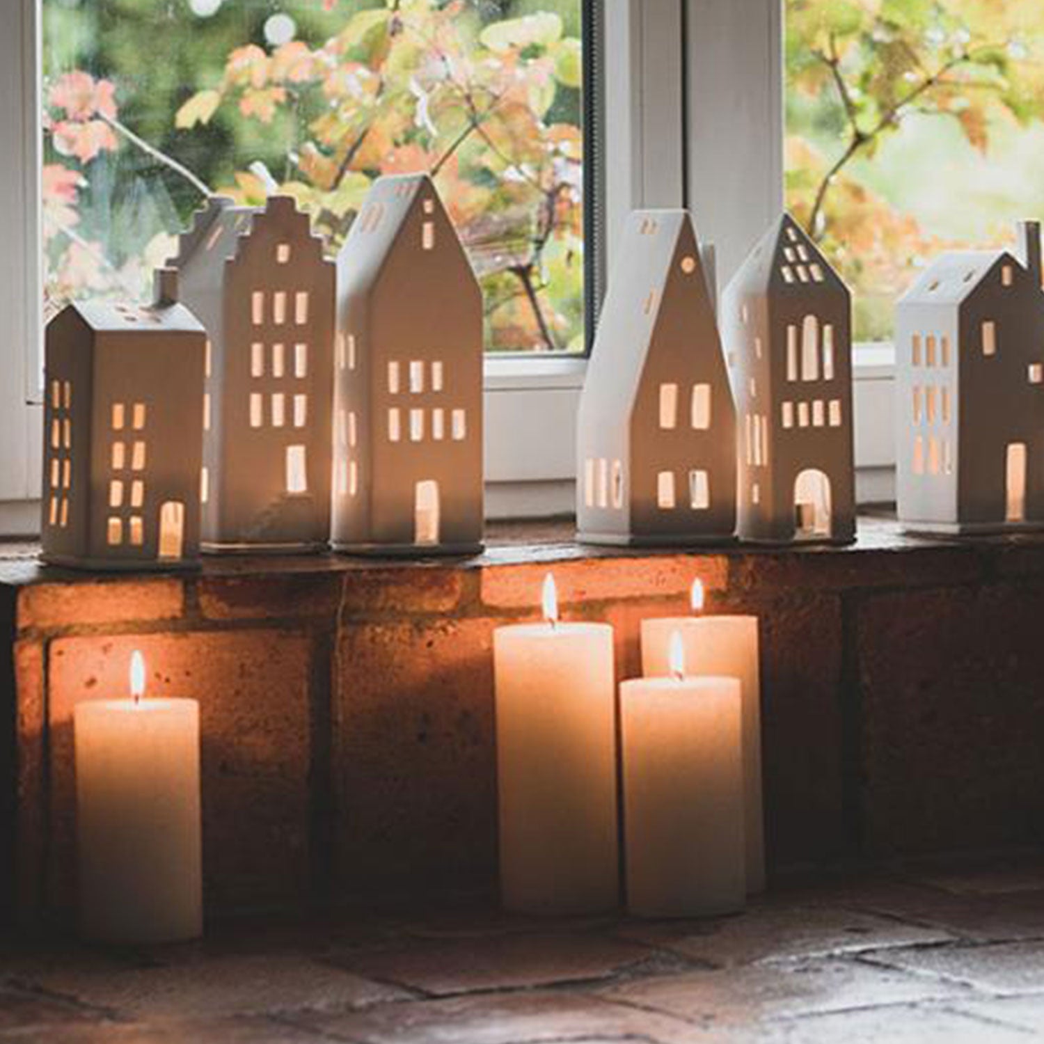 Die Laternen und hohen Kerzen des Kleinen Lichthauses „Spitzdach“ zaubern ein warmes, festliches Licht auf die Fensterbank – perfekt für Weihnachten. Der Blick nach draußen zeigt verschwommenes grünes Laub und schafft eine gemütliche und ruhige Atmosphäre.