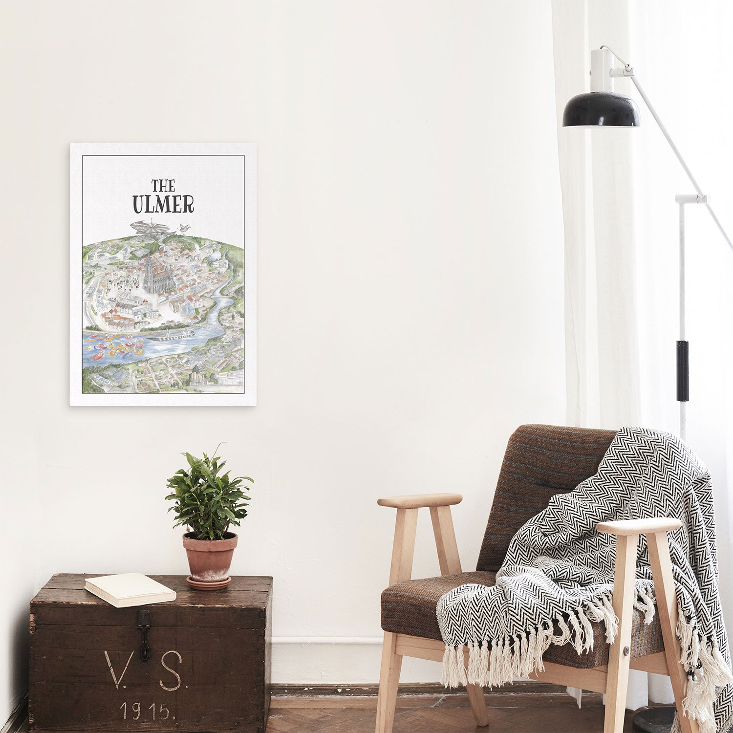 Ein gemütlicher Raum präsentiert ein Kunstdruck-Kartenposter „The Ulmer“, einen minimalistischen Holzstuhl, der mit einer schwarz-weißen Decke bedeckt ist, eine kleine Topfpflanze auf einer Vintage-Truhe und eine moderne Stehlampe, die eine luftige und helle Atmosphäre schaffen.