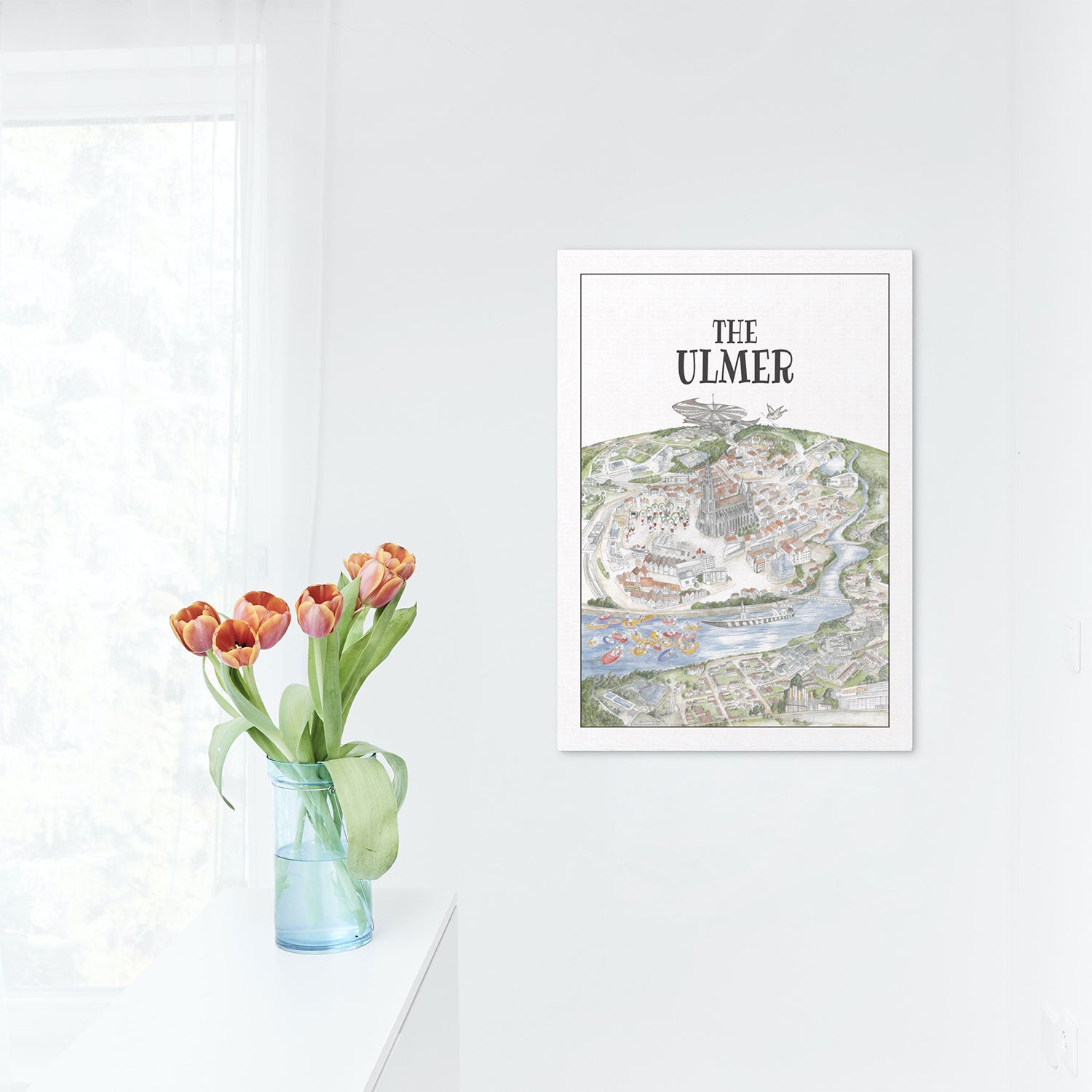 Ein heller, minimalistischer Raum präsentiert eine Vase mit roten und gelben Tulpen auf weißem Untergrund. „The Ulmer“ aus der Kunstdruck-Kollektion hängt gerahmt an der Wand, während sanftes Licht durch einen transparenten Fenstervorhang fällt.