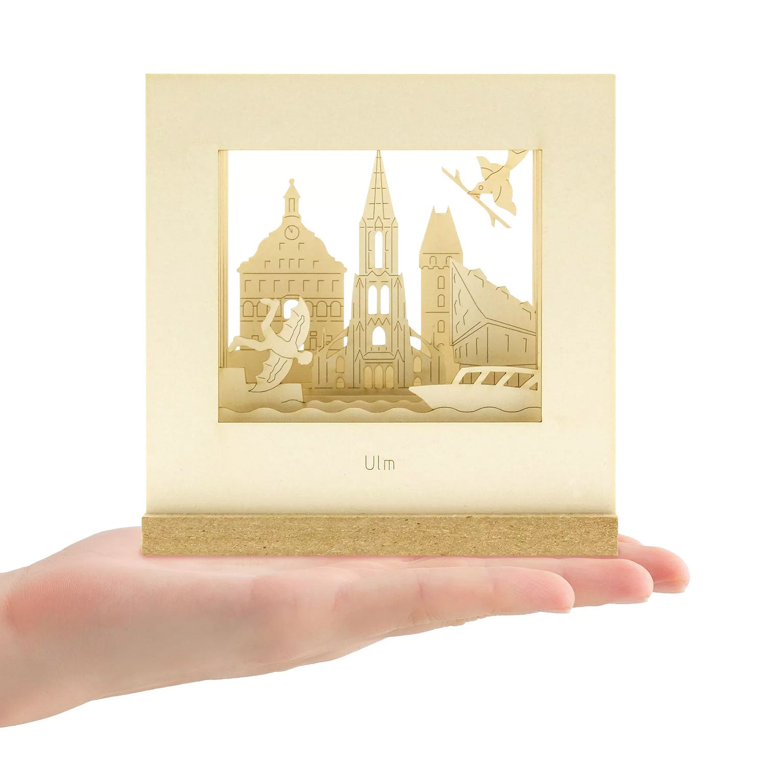 Eine Hand hält die Ulm-SILHOURAMA Silhoubox L, einen dekorativen Holzrahmen mit 3D-Ansichten der Ulmer Dome und Gebäude. Die detailreiche Szenerie im minimalistischen Ständer ist das perfekte Geschenk für Ulm-Fans oder als unvergessliches Souvenir aus Ulm.