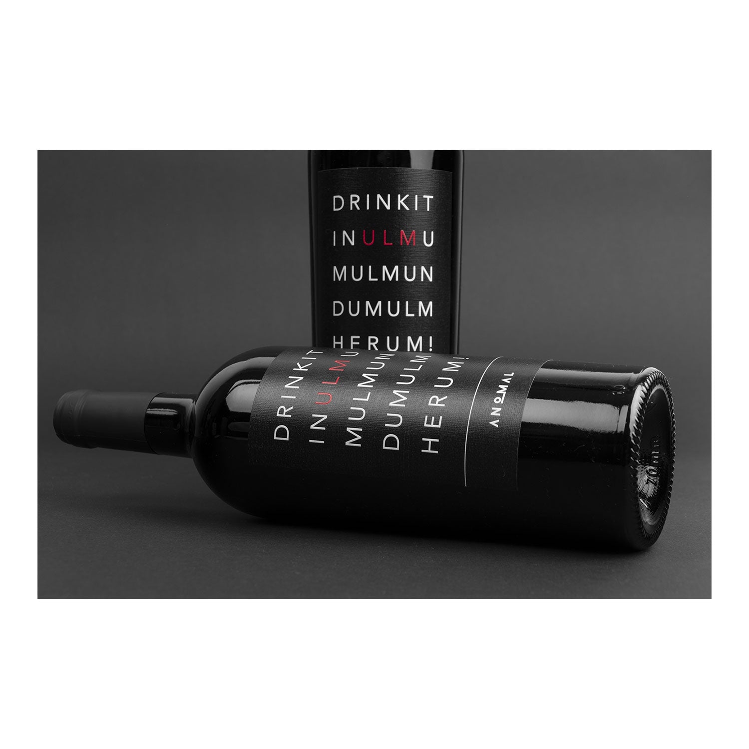 Zwei Anomal-Wein-Flaschen mit dem schwarzen Etikett „DRINK IT IN MULMUL DUMUL HERUM!“ – eine stehend, eine liegend – im Stil „ANOMALI“ vertikal, bieten Ihnen den fesselnden Reiz von 3 Flaschen Wein – Zungenbrecher.
