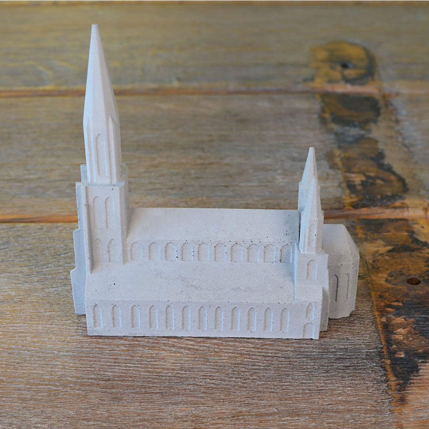 Das Ulmer Münster Beton ist ein Miniaturmodell einer Kathedrale aus grauem Beton mit spitzen Türmen und rechteckigen Fenstern. Es wurde im modernen 3D-Druckverfahren gefertigt und präsentiert sich elegant auf einer Holzoberfläche.