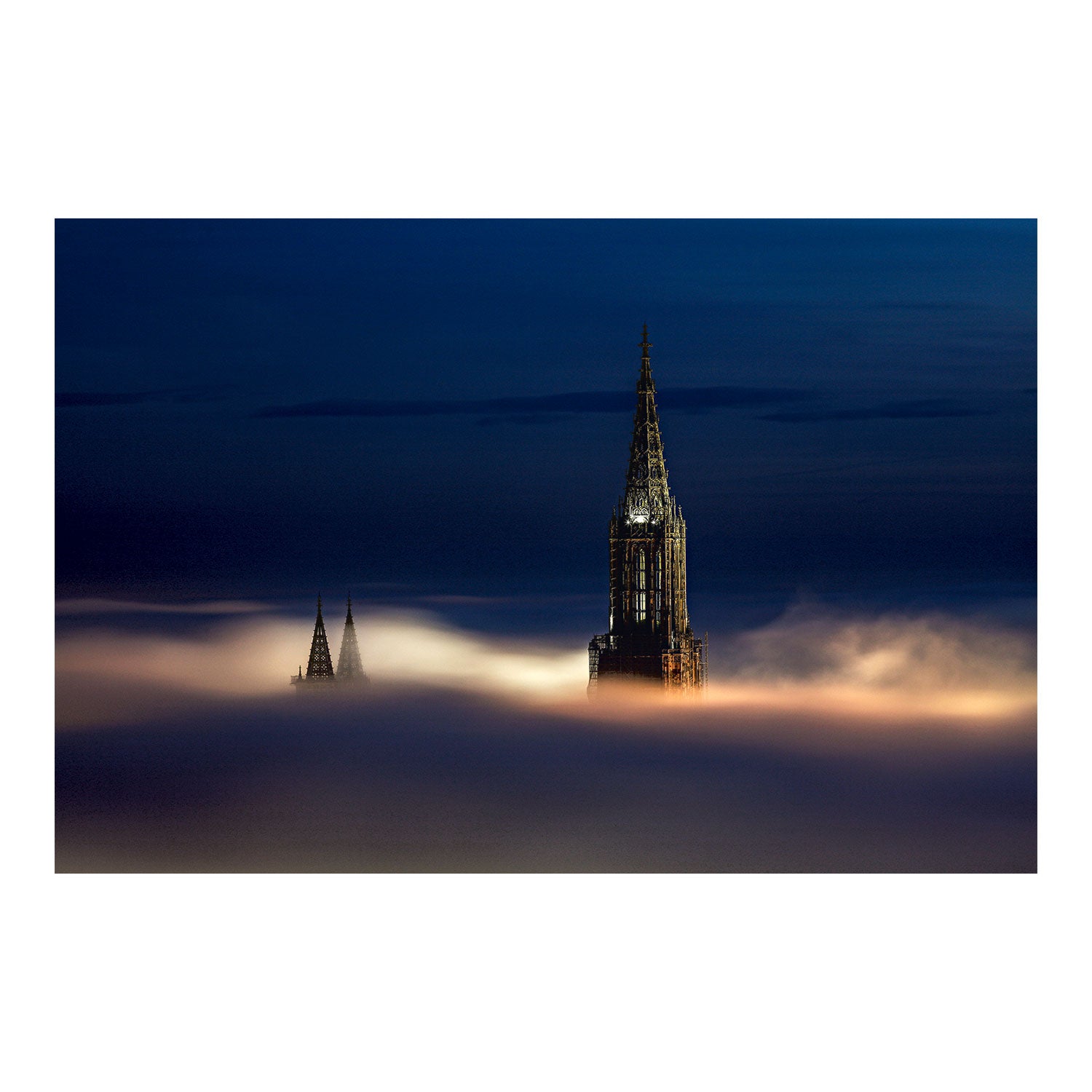 Die große Version von „Ulmer Münsterspitze bei Nacht“ zeigt zwei gotische Kirchtürme, die durch den nächtlichen Nebel ragen. Ihre Beleuchtung kontrastiert mit dem dunklen Himmel und erzeugt eine mystische Atmosphäre. Ideal als Alu-Dibond-Fotodruck-Souvenir aus Ulm.