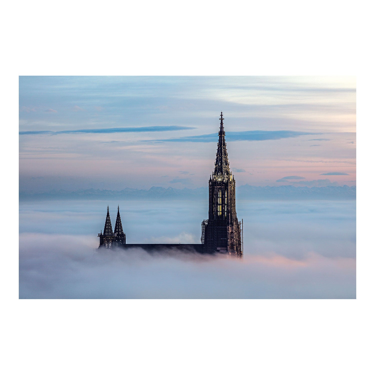 Die „Ulmer Münsterspitze bei Tag – kleine Version“ zeigt einen hohen Turm einer Kathedrale, der sich bei Sonnenaufgang oder Sonnenuntergang über die Wolken erhebt, vor einem pastellfarbenen Himmel und fernen Bergen, und spiegelt die Eleganz gotischer Architektur im ruhigen Fotodruck-Stil wider.