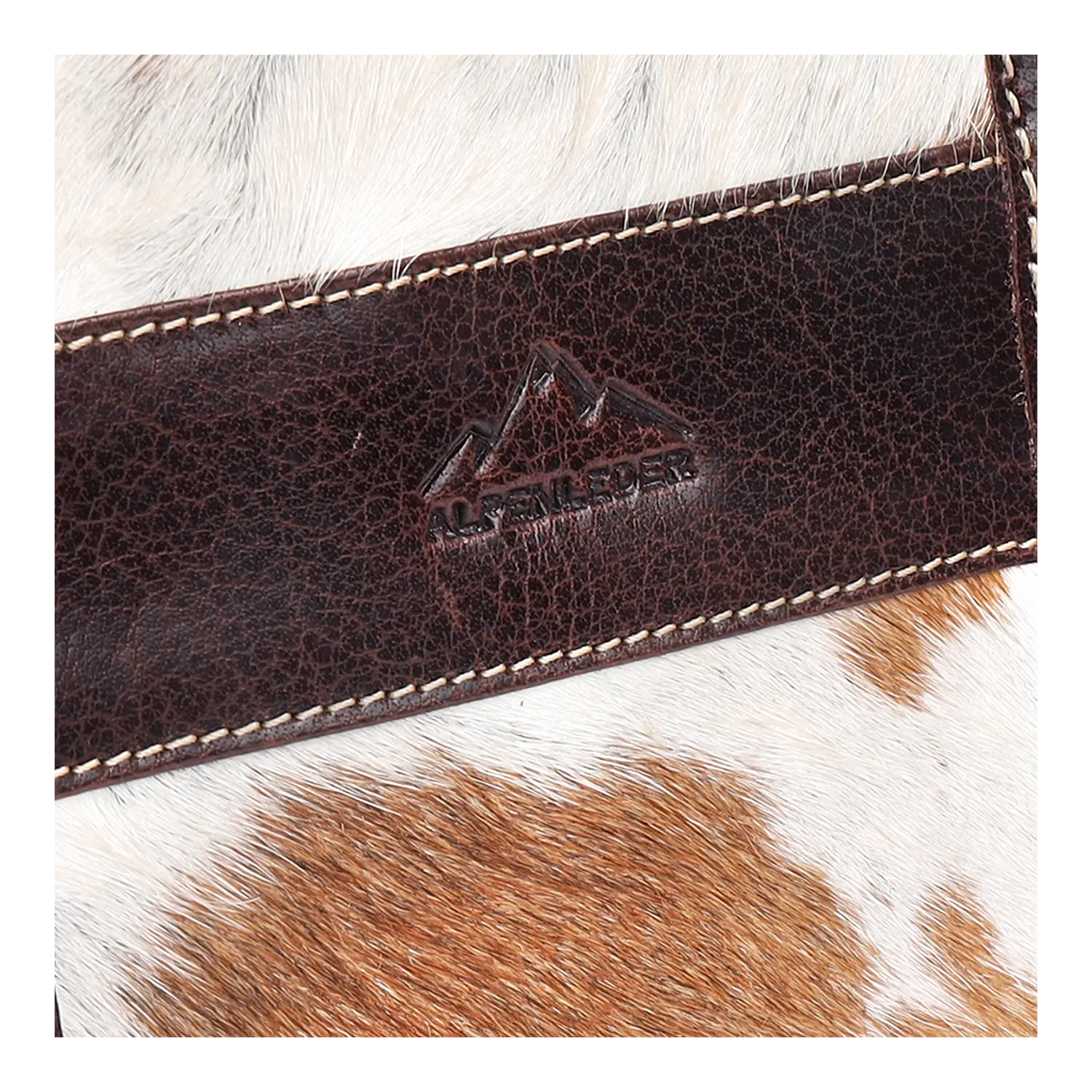 Nahaufnahme eines Alpenleder-Lederpatches mit geprägten Bergen und Text auf einer Oberfläche mit braun-weißem Kuhfell-Muster. Das Leder aus der Umhängetasche SPRUCHBEITEL-Kollektion weist detaillierte Nähte an den Rändern auf.