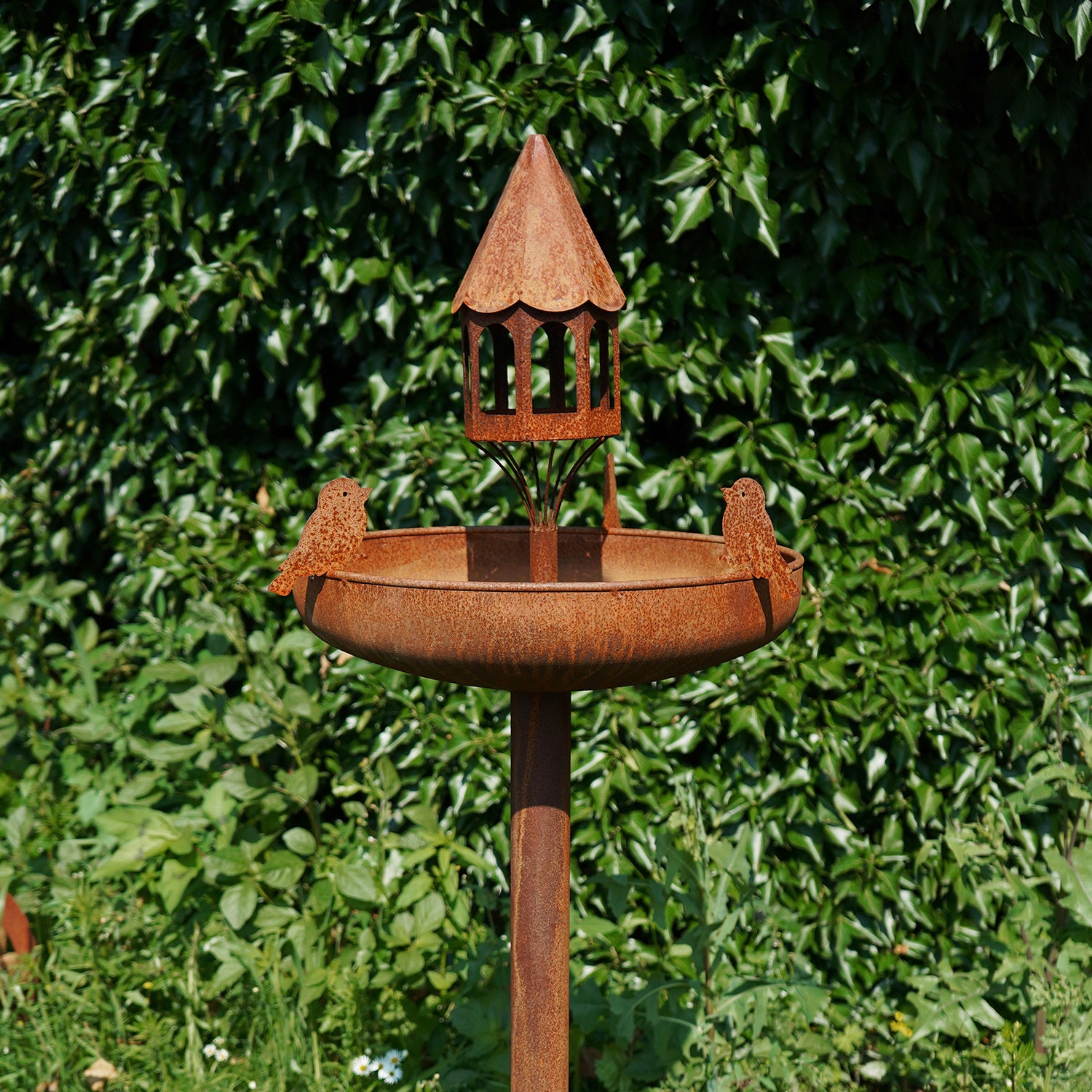 Eine Vogeltränke aus Edelrost mit kleinem Dach und zwei Vogelsilhouetten wertet einen üppigen Garten auf. Dieses rustikale Vogelbad hebt sich vom dichten grünen Laub ab und erinnert an den Charme des Bayerischen Waldes.