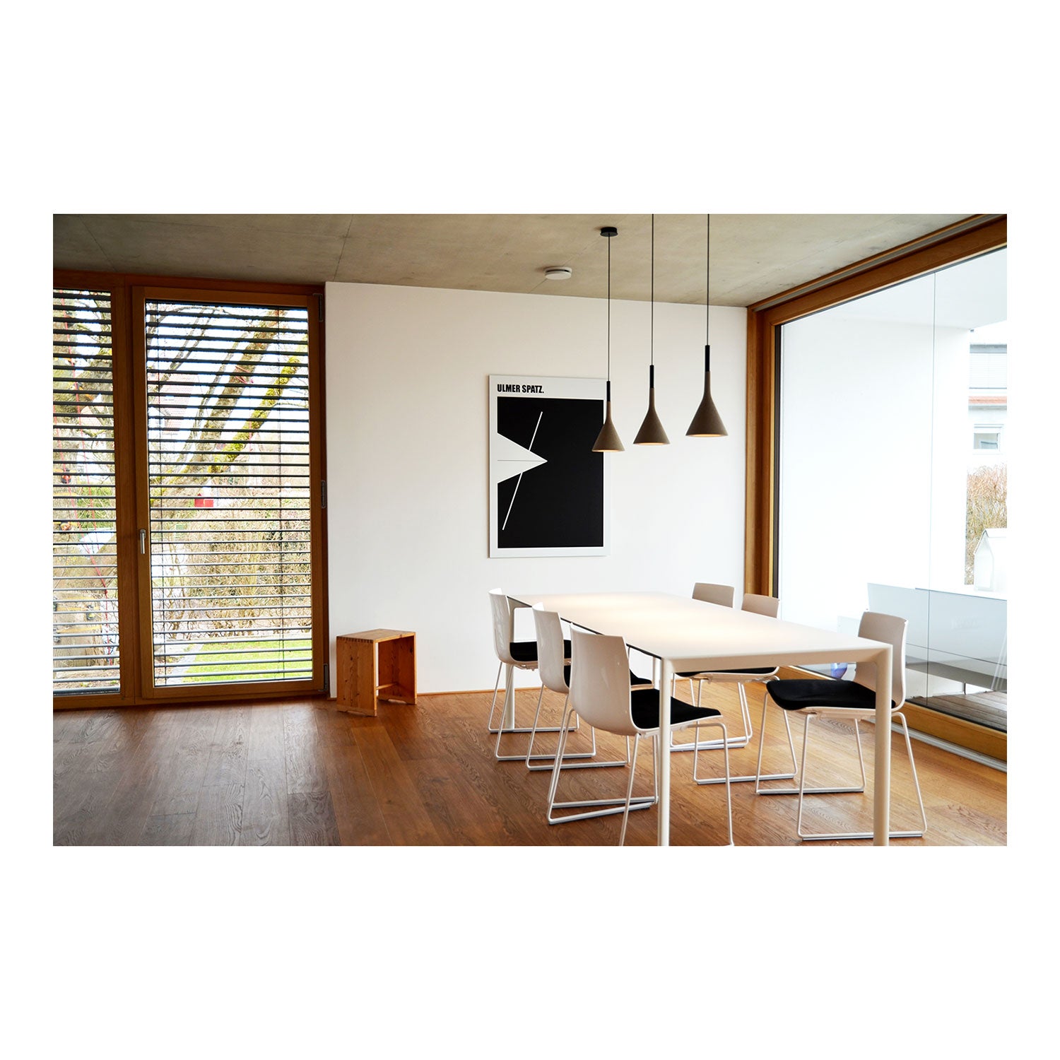 Ein modernes Esszimmer verfügt über einen rechteckigen Tisch, sechs weiße Stühle, drei Hängelampen, große Fenster mit Holzrahmen, minimalistisches Dekor und das Wandbild „Ulmer Schachtel“ – ein abstraktes Kunstwerk in Schwarzweiß – an der Wand.