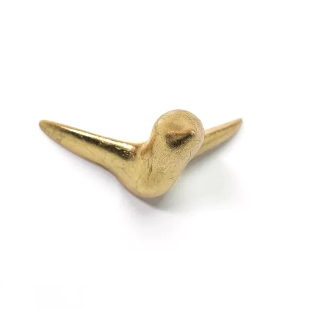 Auf weißem Hintergrund ist eine kleine goldfarbene abstrakte Skulptur mit dem Namen „Wandskulptur Mini Vogel Gold“ zu sehen, die einem Keramikvogel mit ausgebreiteten Flügeln ähnelt.