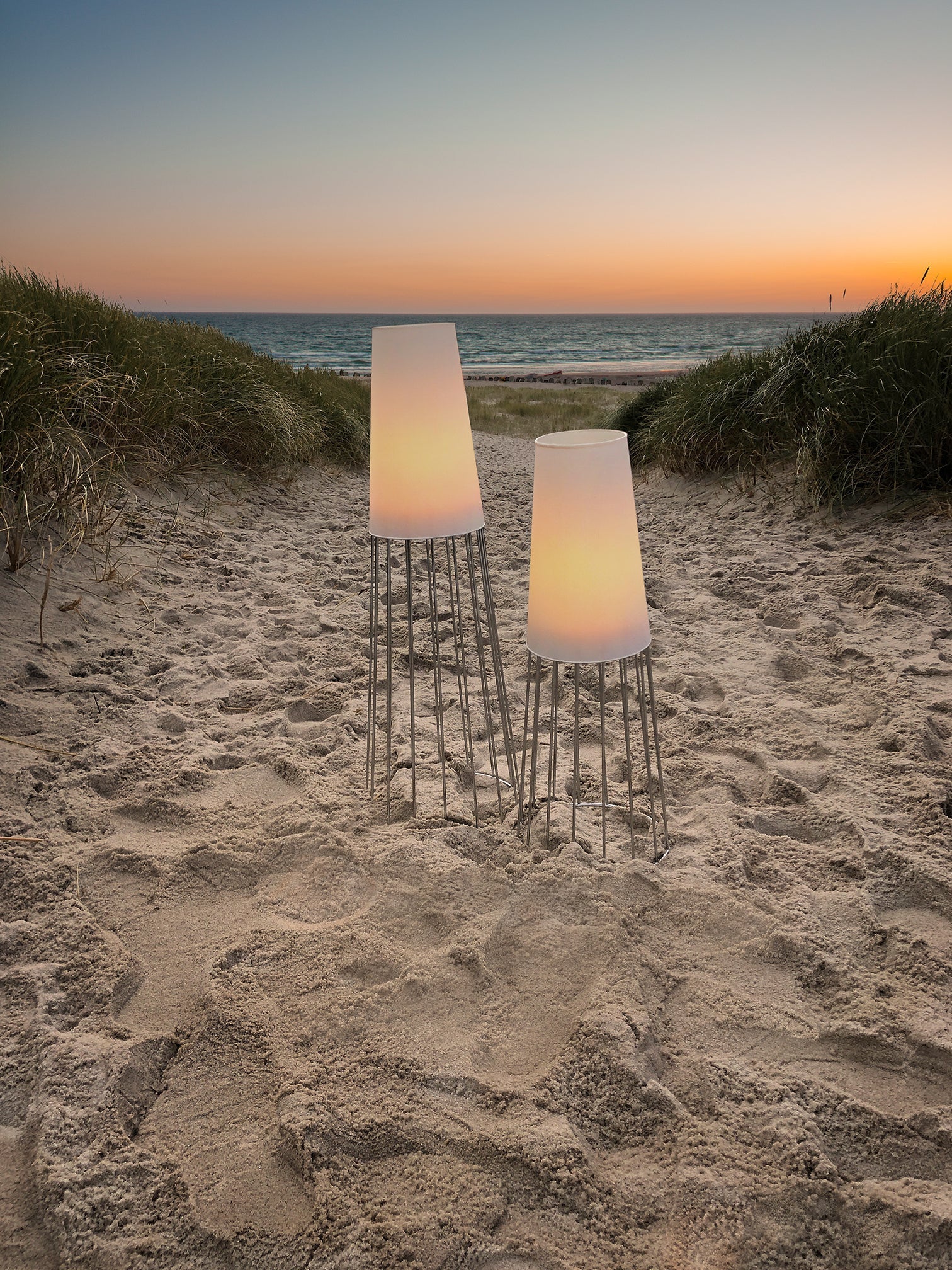 Zwei leuchtende Lampen mit Lichteffekten beleuchten bei Sonnenuntergang einen Sandstrandweg, der von Grasdünen gesäumt ist und im Hintergrund das Meer zeigt – eine ruhige Szene, die an ein Gartenheiligtum erinnert.