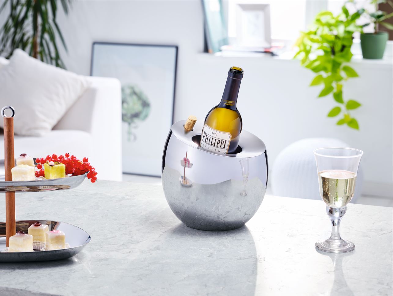 Eine Weinflasche in einem Edelstahl-Weinkühler auf einem Marmortisch, daneben ein Glas Wein, ein Korkenhalter und ein Desserttablett.