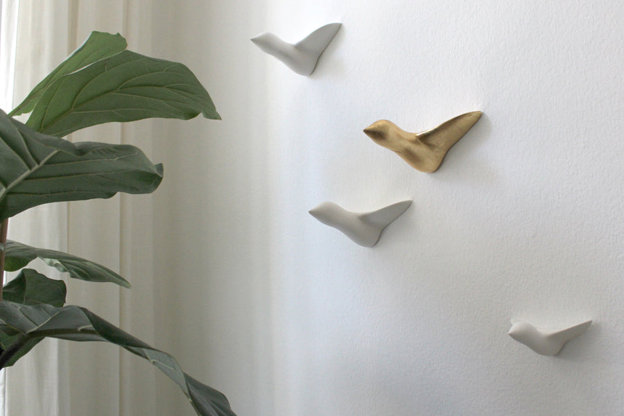 Die Wandskulptur „kleiner Vogelschwarm“ zeigt vier minimalistische, weiß-goldene Keramikvögel im Flug und verkörpert funktionales Design. Eine üppige grüne Pflanze links setzt vor hellem Hintergrund einen natürlichen Akzent.