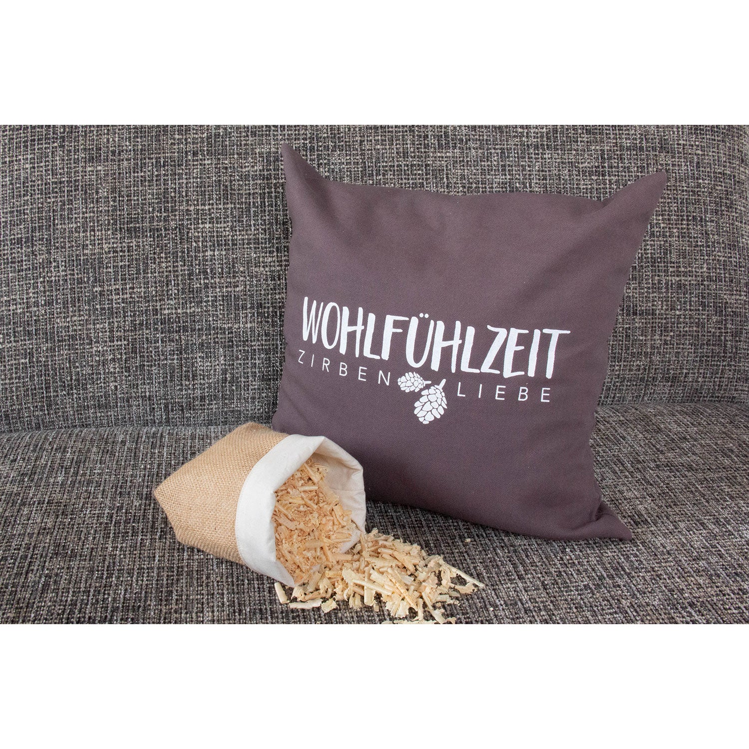 Ein „Kissen Wohlfühlzeit“-Kissen mit dem Text „Wohlfühlzeit Zirbenliebe“ und Tannenzapfengrafiken auf strukturiertem grauem Stoff mit dezentem Zirbenduft-Aroma liegt neben einem Leinensack, aus dem duftende Zirbenholzflocken strömen.