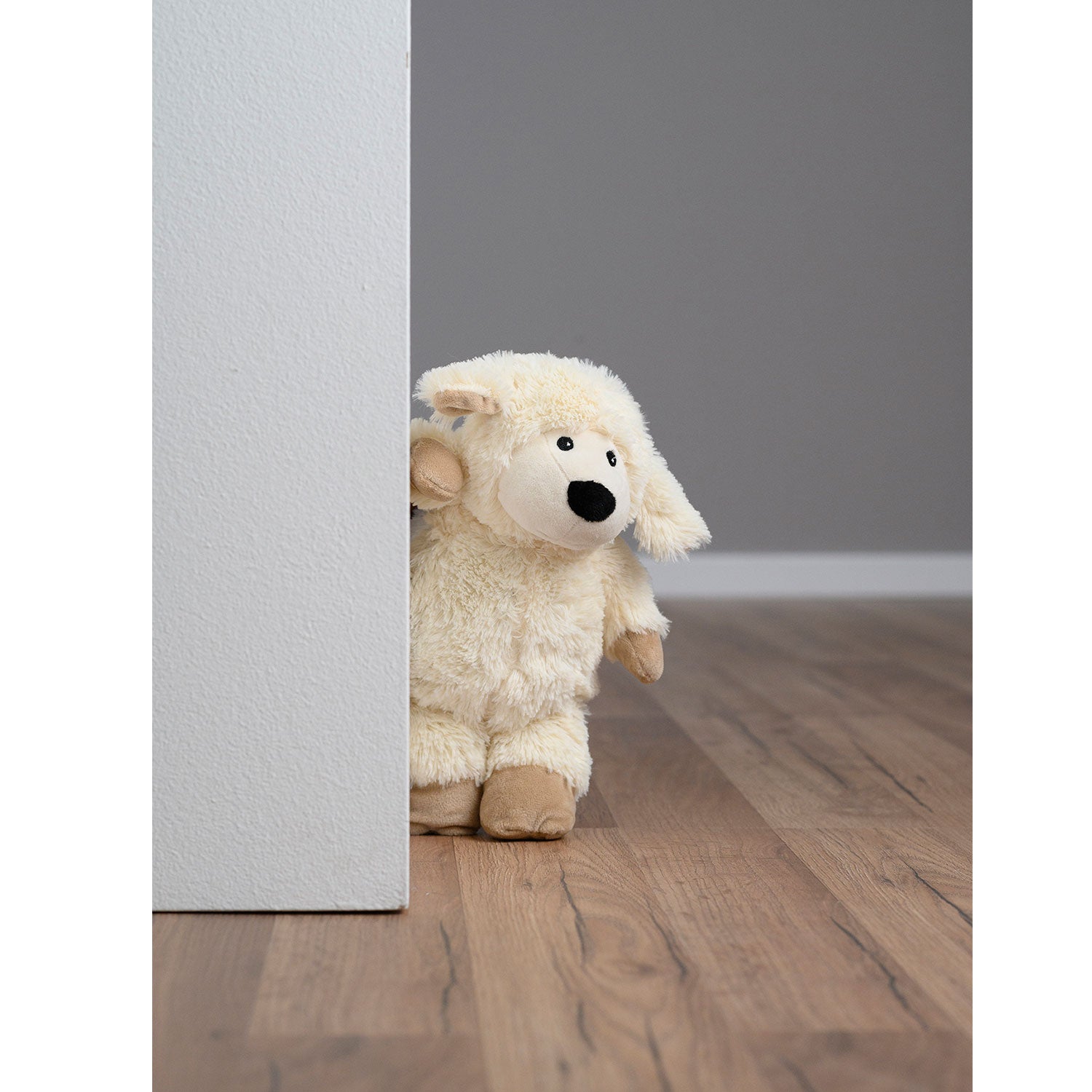 Ein Wärmetier, Schaf Lavendi, lugt hinter einer weißen Wand in einem Raum mit Holzboden hervor. Sein neugieriger Ausdruck und seine weiche Textur heben sich vom neutralen Hintergrund ab und verleihen ihm eine launische Note und Wärme.